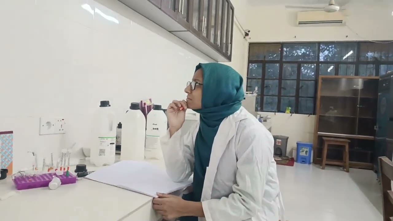 A researcher in a lab coat and headscarf sitting at a white bench, pen in hand, looking up from an open paper notebook, with reagent bottles and a rack of tubes alongside and lab shelving behind.